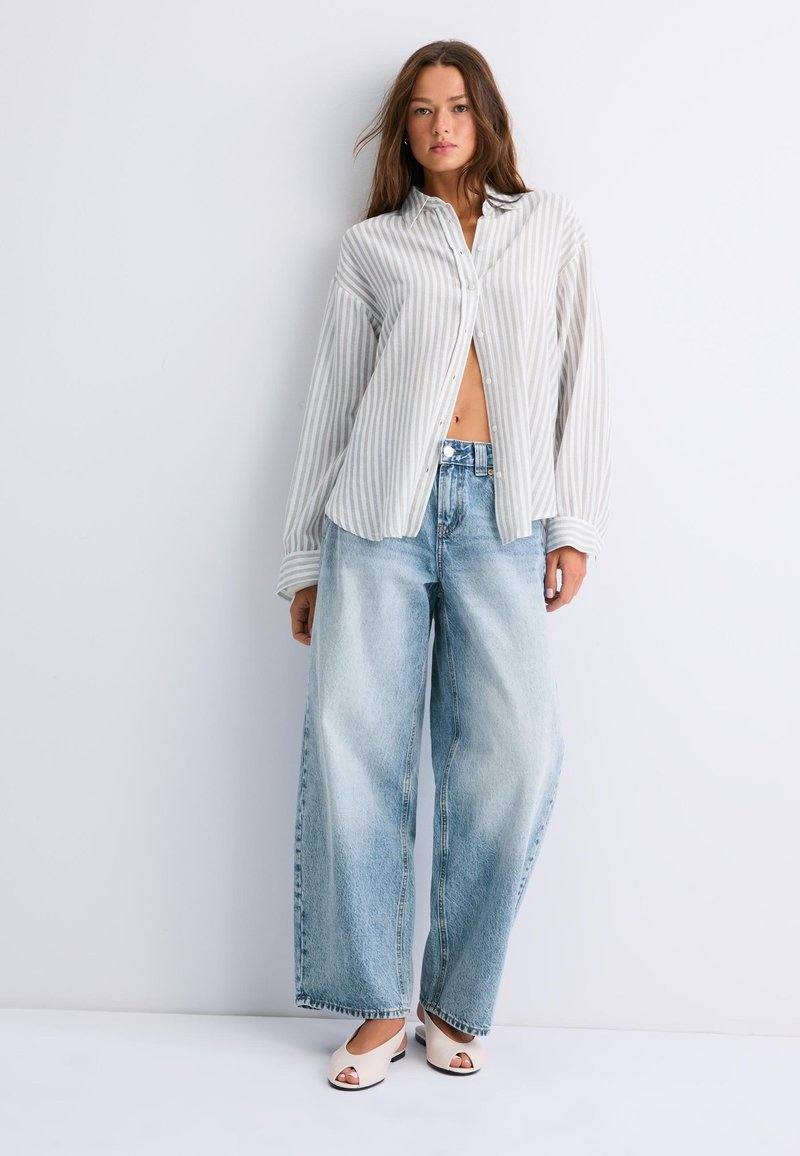 Femme portant une chemise ample blanche et grise rayée, un jean large bleu clair et des chaussures blanches à bout ouvert, debout devant un fond blanc uni.