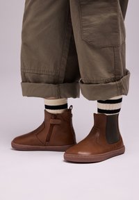 Botas de cuero marrón hasta el tobillo con cremalleras laterales y detalles elásticos, combinadas con pantalones verde oliva arremangados y calcetines a rayas en blanco y negro.