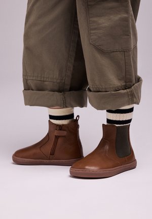 Bottines en cuir marron avec des zips latéraux et des accents en élastique, associées à un pantalon olive roulé et des chaussettes rayées noir et blanc.