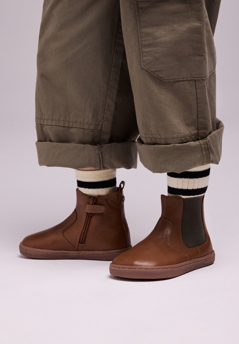 Botas de cuero marrón hasta el tobillo con cremalleras laterales y detalles elásticos, combinadas con pantalones verde oliva arremangados y calcetines a rayas en blanco y negro.