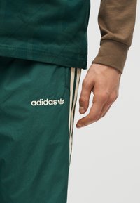 Ανδρικό χέρι κοντά σε πράσινο παντελόνι Adidas με άσπρες ρίγες και λογότυπο, φορώντας καφέ μακρυμάνικη μπλούζα σε απλό φόντο.
