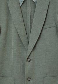 Blazer verde testurizzato con revers a lancia, due bottoni anteriori e taschino sul petto. Interno foderato con tessuto azzurro chiaro visibile al colletto.