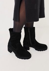 Botines negros de ante, con cierre de cremallera, parte superior texturizada y suelas de goma robustas. Acentos de doble costura a lo largo de los lados.