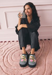 Pantofole slip-on a righe bianche e nere con ricamo floreale recante la scritta "NO RISK NO MAGIC". Texture morbida su un tappeto rosa a onde.