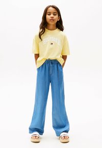 Żółta bawełniana koszulka z logo "HILFIGER" w kolorze białym, w połączeniu z luźnymi niebieskimi dżinsami i białymi sandałami. Gładkie tło.