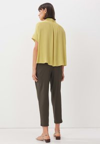 Blusa a maniche corte di un leggero giallo chiaro con piega sul retro, abbinata a pantaloni verde scuro e sandali minimalisti in argento.