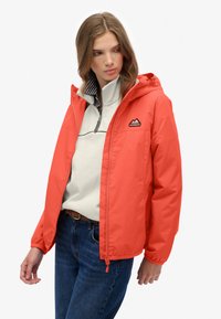 Ung kvinde iført en rød hættejakke over en lys, lynlåstrøje og blå jeans, der ser mod venstre mod en hvid baggrund.