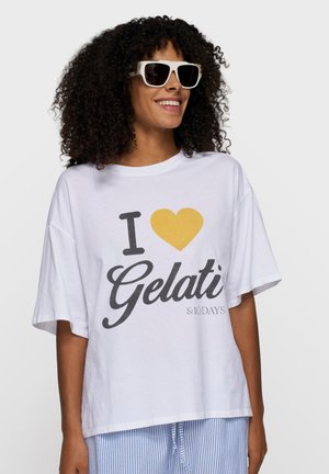 Donna sorridente con capelli ricci, occhiali da sole bianchi, maglietta bianca oversize con la scritta "I love Gelati" e pantaloni a righe blu.