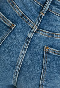 Blå denimjeans med synliga bakfickor och framträdande orange söm i en närbild.