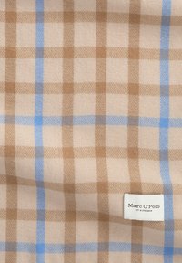 Tissu beige avec un grand motif à carreaux marron et bleu clair, et une étiquette de marque blanche indiquant "Marc O’Polo EST. À STOCKHOLM"