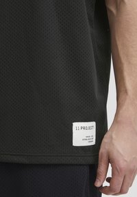 Sort mesh T-shirt med et perforeret design. Inkluderer et hvidt stofmærke ved kanten med mærkenavn og oplysninger.