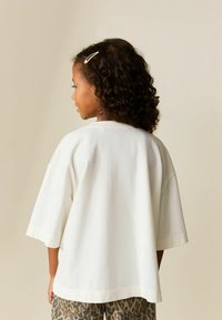 Valge liibuvate mõõtmetega t-särk pehme kangaga, lühikeste varrukatega, vabalt langev lõige, kombineeritud pruunide leopardimustriliste pükstega, vaadatuna tagant.