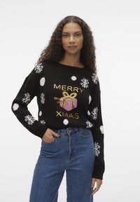 Sort genser med hvite snøfnugg og prikkete mønster, med teksten "MERRY XMAS" i gull og en rosa gaveeske-illustrasjon. Strikket tekstur, komfortabel passform.