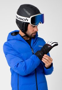 Skihandskar i svart och vitt, med texturerad tyg, elastiska manschetter och en logotyp. Bärs av en person i en blå pufferjacka och hjälm.