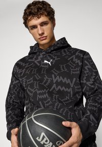Sort hættejakke med et abstrakt gråt mønster, der har en frontlomme og Puma-logo. Personen holder en sort basketball med sølvdetaljer.