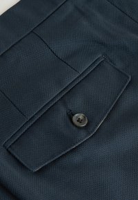 Gros plan sur un tissu bleu foncé texturé avec une poche à rabat maintenue par un seul bouton sombre.