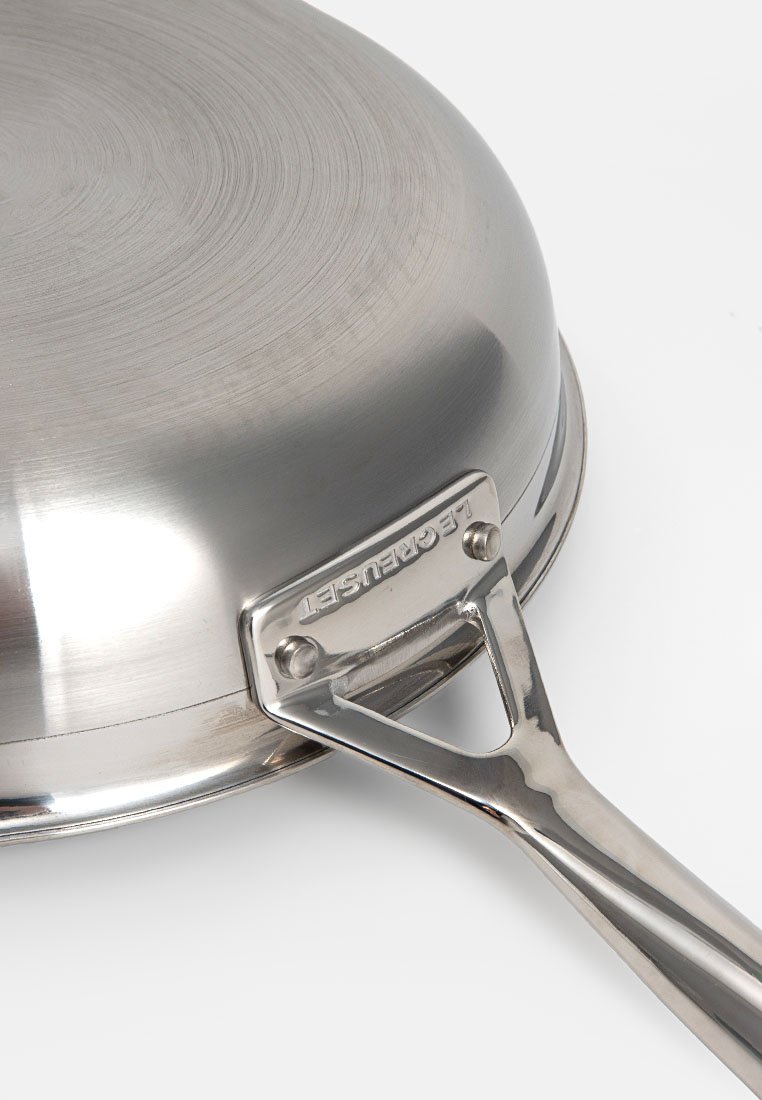 Edelstahl-Bratpfanne von unten gezeigt, mit schrägem Griff, der mit Nieten befestigt ist, Markenname "Le Creuset" am Griffansatz eingraviert.
