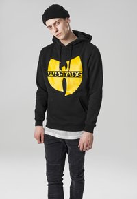 Sudadera negra con el logo amarillo de Wu-Tang en el centro. Presenta un bolsillo tipo canguro, capucha con cordón y puños y dobladillo acanalados. Se lleva con jeans negros.