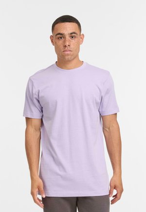 Homme aux cheveux courts et aux taches de rousseur, portant un T-shirt simple de couleur violet clair et un pantalon sombre, se tenant devant un fond blanc.
