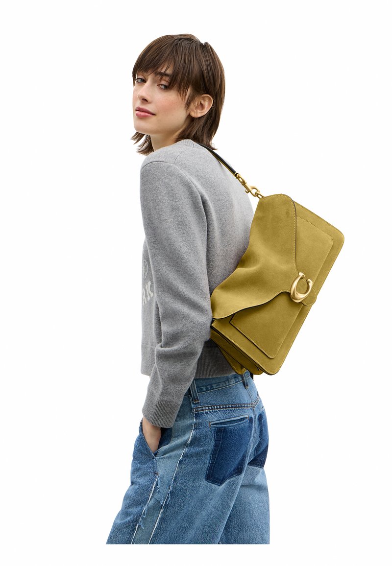 Femme en pull gris et jean patchwork portant un grand sac à bandoulière moutarde avec une boucle dorée, regardant par-dessus son épaule.