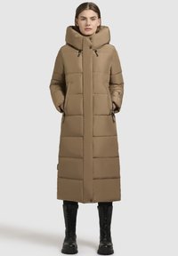 Dlhý hnedý puff coat s vysokým golierom, horizontálnym prešívaním, bočnými vreckami a rovným lemom; nosený s čiernymi čižmami po kolená.