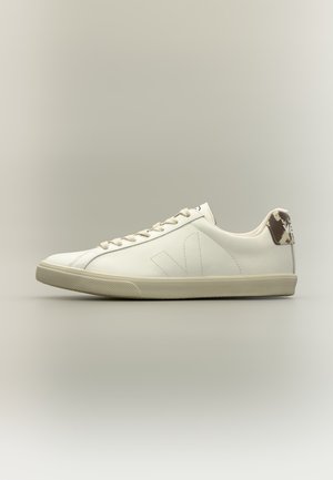 Hvid lavtopet sneakers med beige sål, snørebånd og en brun mønstret hælflig mod en neutral baggrund.