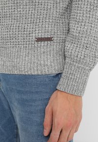 Pull gris en tricot avec texture côtelée, avec une étiquette marron "PIER-ONE" sur le côté. Associé à un jean en denim bleu clair.