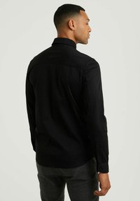 Camisa negra de manga larga con cuello alto, textura suave, puño abotonado y corte entallado. Detalles de costura visibles en los hombros.