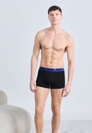 Homme torse nu debout à l'intérieur portant un boxer Puma noir avec une ceinture bleue, expression neutre, fond gris clair, et une chaise moderne à proximité.