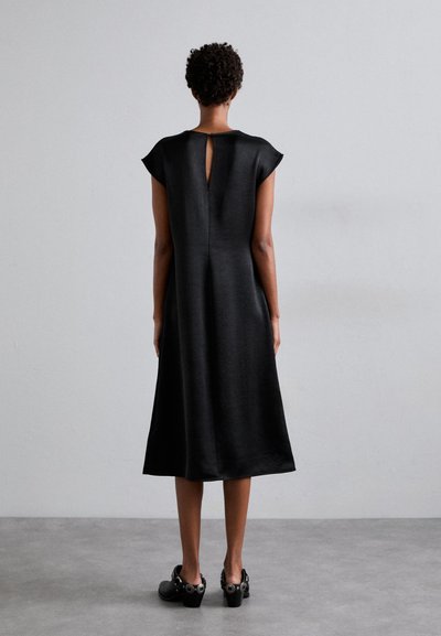 Black, knee-length dress with cap sleeves, a keyhole back, and a smooth, textured fabric. Paired with clogs featuring metal accents.