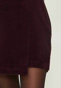 Korte corduroy rok in diep bordeaux, met een zijslit en subtiele ribbelstructuur. De zoom is afgewerkt met schone randen. Dunne zwarte panty's eronder.