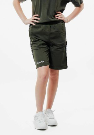 Una persona in piedi con le mani sui fianchi indossa pantaloncini sportivi verde scuro e scarpe da ginnastica bianche su sfondo bianco.