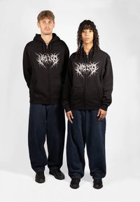 Svart hettejakke med glidelås har et sølvfarget grafisk design. Kombinert med løstsittende denimjeans og svarte sko. En enkel bakgrunn.