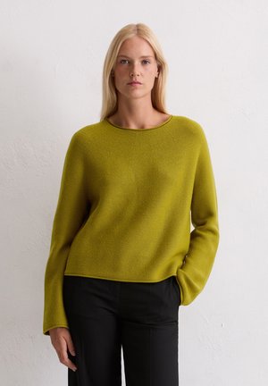 Blonde vrouw die tegen een witte muur staat in een mosgroene gebreide trui en zwarte broek, met één hand in haar zak.