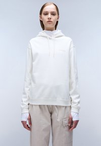 Felpa bianca con cappuccio e un logo discreto sul petto. Il tessuto è liscio, abbinato a pantaloni beige chiaro.