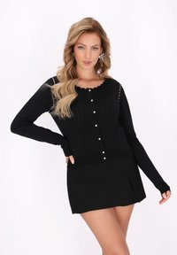 Cárdigan negro de punto acanalado con escote de picos, botones frontales y mangas largas; combinado con una mini falda negra a juego.
