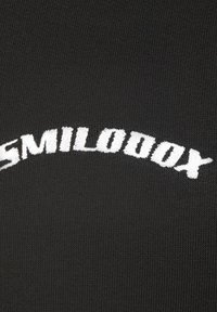 Tissu en tricot noir avec un texte brodé blanc en courbe lisant "SMILODOXX" sur le devant, présentant une texture fine et des coutures régulières.