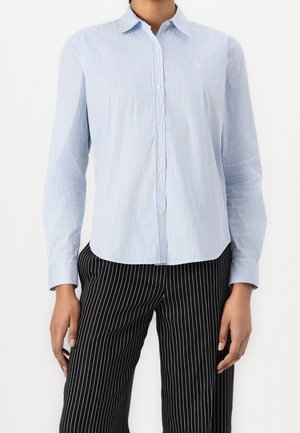 Osoba oblečená v svetle modré a bílo pruhované košili na knoflíky a černé kalhoty s bílými proužky, stojící na jednobarevném pozadí.