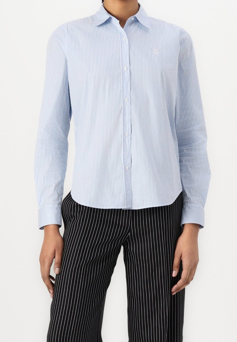 Osoba nosząca jasnoniebieską i białą koszulę w cienkie prążki zapinaną na guziki oraz czarne spodnie w białe prążki, stojąca na tle jednolitego tła.