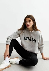 Felpa grigia con il testo "MIAMI", pantaloni jogger neri e sneakers bianche. Outfit casual con una vestibilità rilassata e morbida texture in cotone.