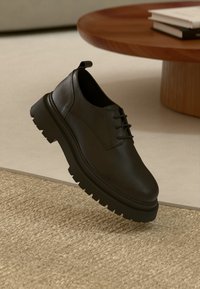Black leather shoe with a textured surface, rounded toe, and lace-up design. Features a thick, rugged sole with deep grooves for traction.