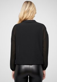Blusa nera con maniche trasparenti, caratterizzata da un collo a camicia e una vestibilità morbida. Tessuto liscia, che termina in vita.