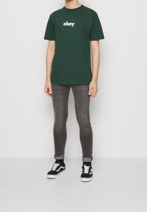 T-shirt en coton vert foncé avec le texte "obey" en blanc, associé à un jean skinny gris et des baskets noir et blanc. Tenue simple et décontractée.