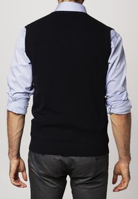 Marinblå stickad väst med ribbad nederkant, buren över en ljusblå rutmönstrad skjorta. Skjortans ärmar är uppvikta, vilket avslöjar en slät tygtextur.