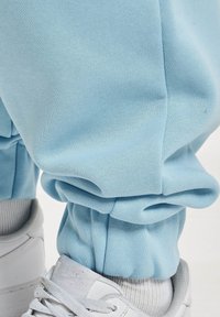 Pantaloni de tip jogger, de culoare albastru deschis, confecționați dintr-un material moale, cu manșete elastice, asortați cu adidași albi cu accente gri și șireturi texturate.