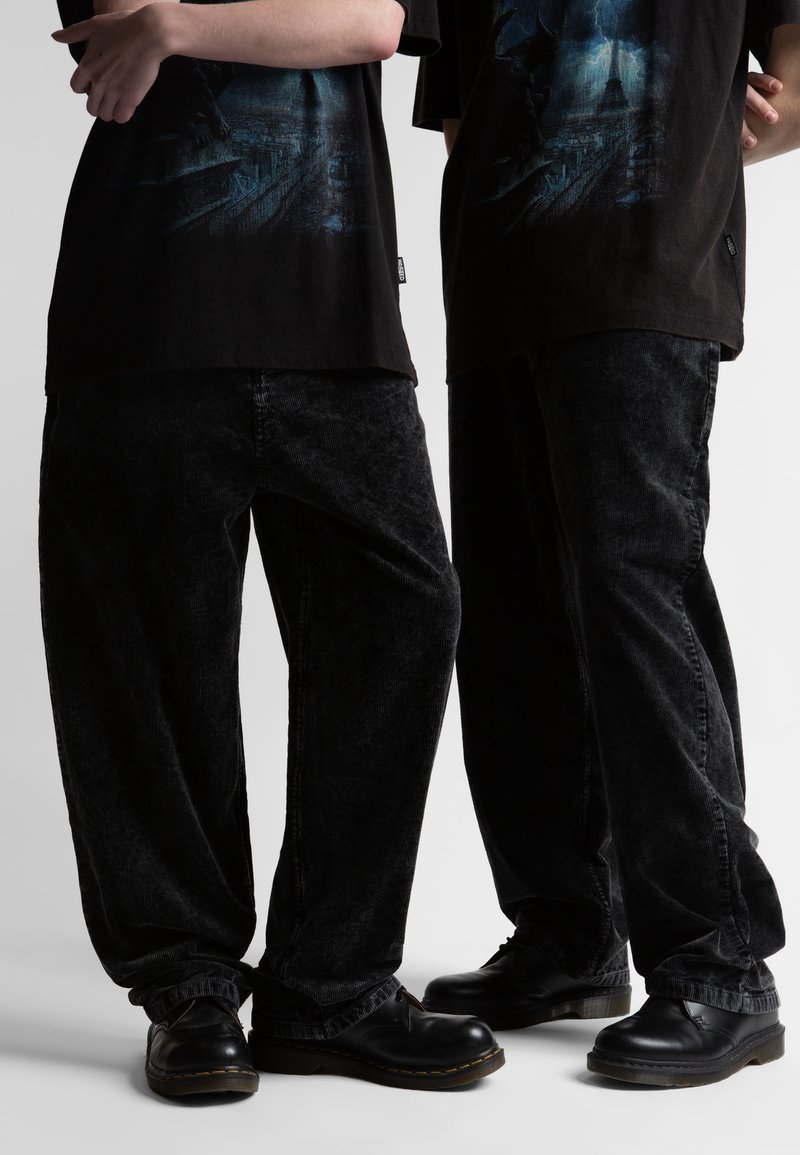 Δύο άτομα φορούν μαύρα γραφικά μπλουζάκια, φαρδιά μαύρα παντελόνια κοτλέ και μαύρα δερμάτινα παπούτσια, στέκονται με σταυρωμένα τα χέρια.