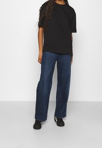 Person med långt flätat hår iklädd en lös svart t-shirt, vida blå jeans och svarta sneakers mot en enkel vit bakgrund.