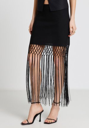 Gonna nera al ginocchio con orlo a frange annodate, abbinata a sandali neri con tacco alto e anelli dorati alle dita.