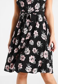 Sukienka w kwiatowy wzór na czarnym tle w groszki, z różowymi i białymi kwiatami, z paskiem w talii oraz zwiewną spódnicą.