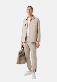 Veste beige avec deux poches avant, associée à un pantalon assorti et des baskets blanches. Sac brun avec une bandoulière et des accents latéraux.